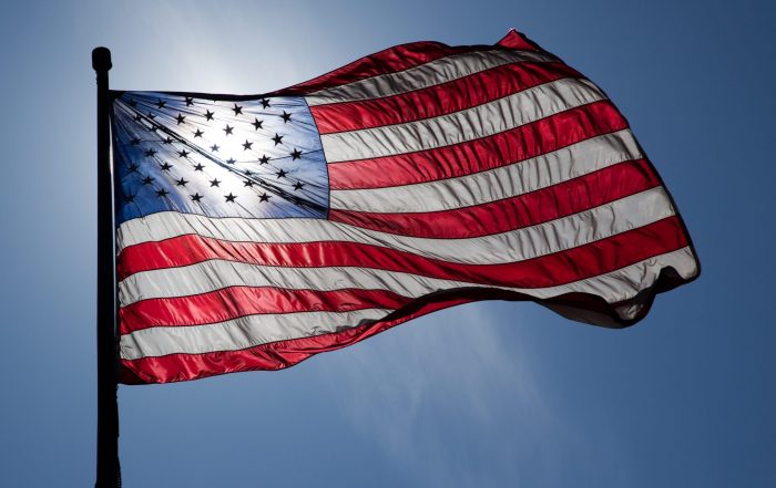 An American flag. The sun is behind it, shining through the stars on the flag.