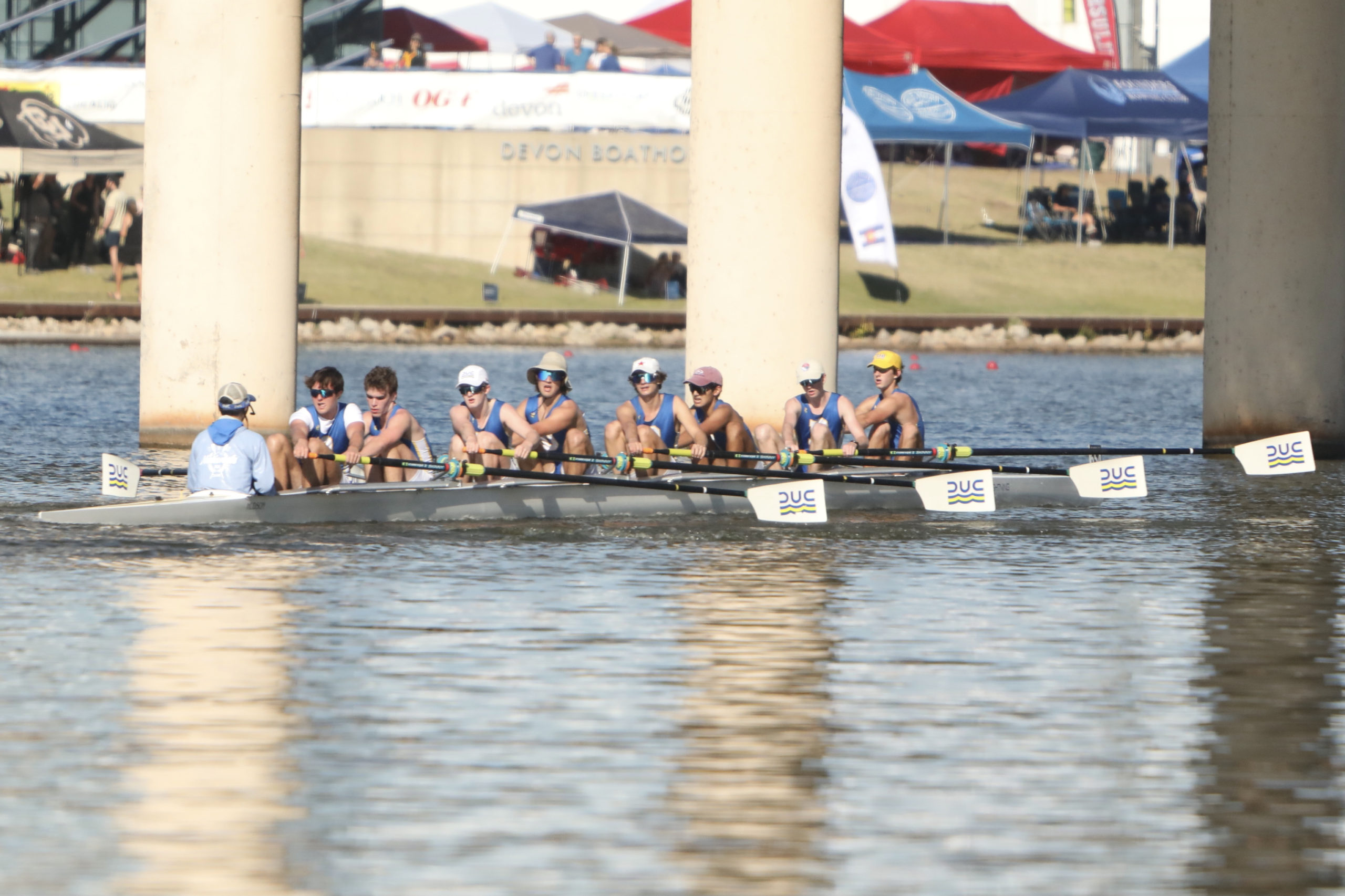 Dallas United Crew varsity boys rowing. There are eight of them, and the blades of their oars are in the air as they prepare to take a stroke.