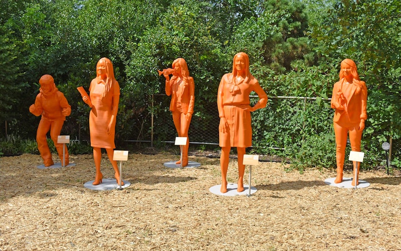 Orange statues of women with trees in the background. The statues are part of the #ifthenshecan series.