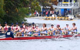 a rowing boat of eight rowers and one coxswain rows past a rival boat. they are preparing to turn a corner on the river. another crew is ahead of them.