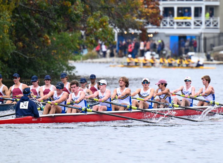 a rowing boat of eight rowers and one coxswain rows past a rival boat. they are preparing to turn a corner on the river. another crew is ahead of them.